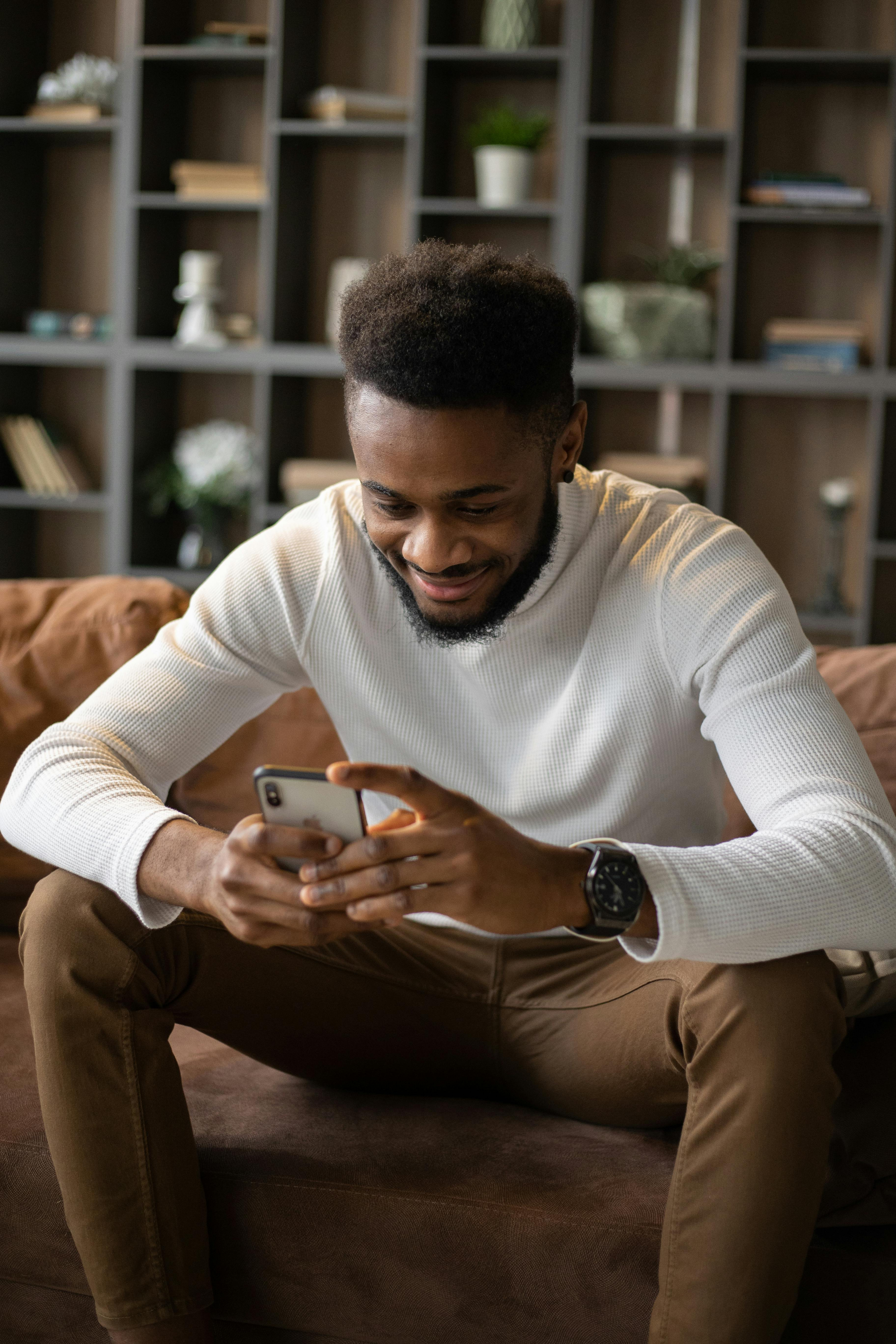 A young man sitting on a brown leather sofa in a warm living room, smiling as he looks at his phone