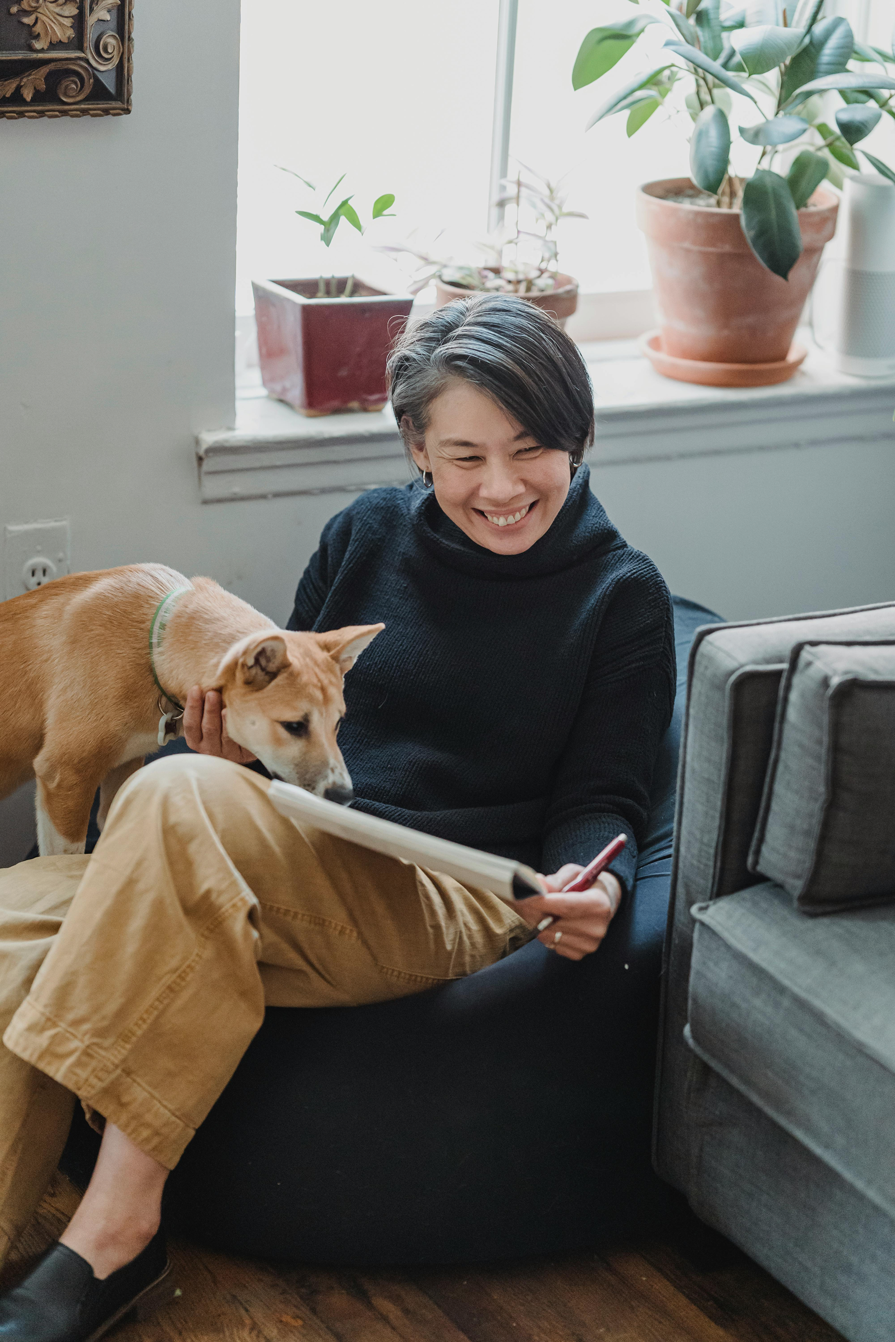 A woman smiling while her golden dog rests against her on a gray sofa, with potted plants on a windowsill behind